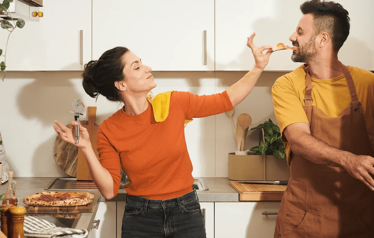 Zwei Menschen probieren Pizza in einer Wohnküche